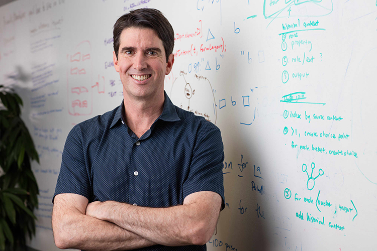 Portrait of Adam Cheyer, Creator of Siri standing in front of a whiteboard with text.