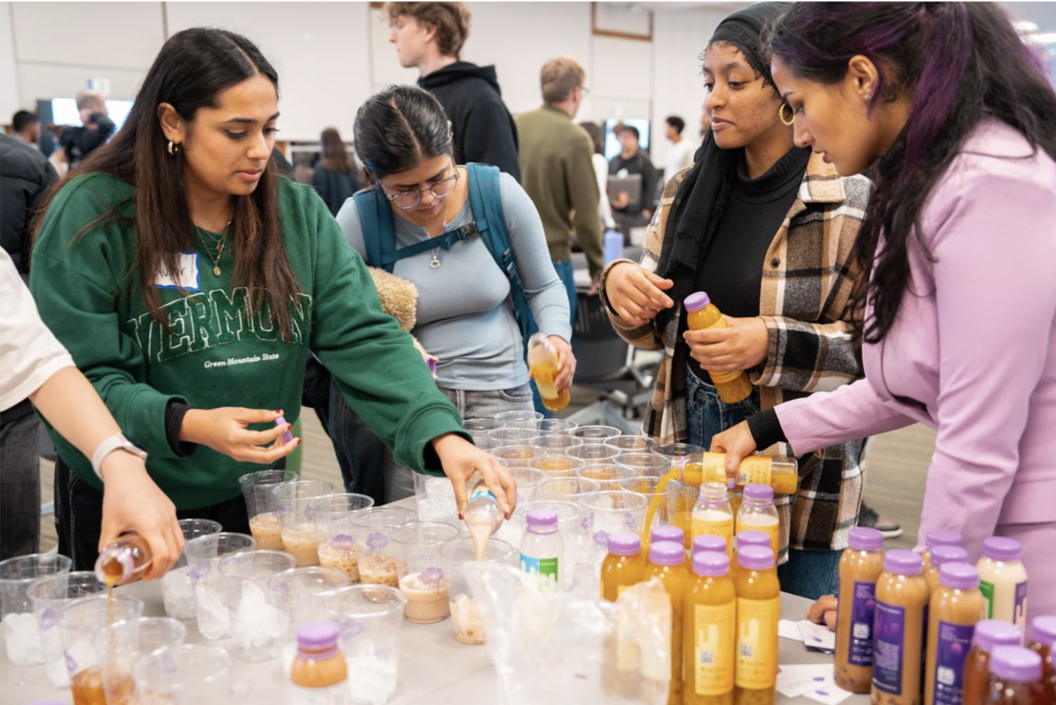 Kashish Juneja pours a small bottle of orange AURA drink into one of many empty clear glasses set up on a table. Four students watch while pouring their own AURA drinks into glasses.