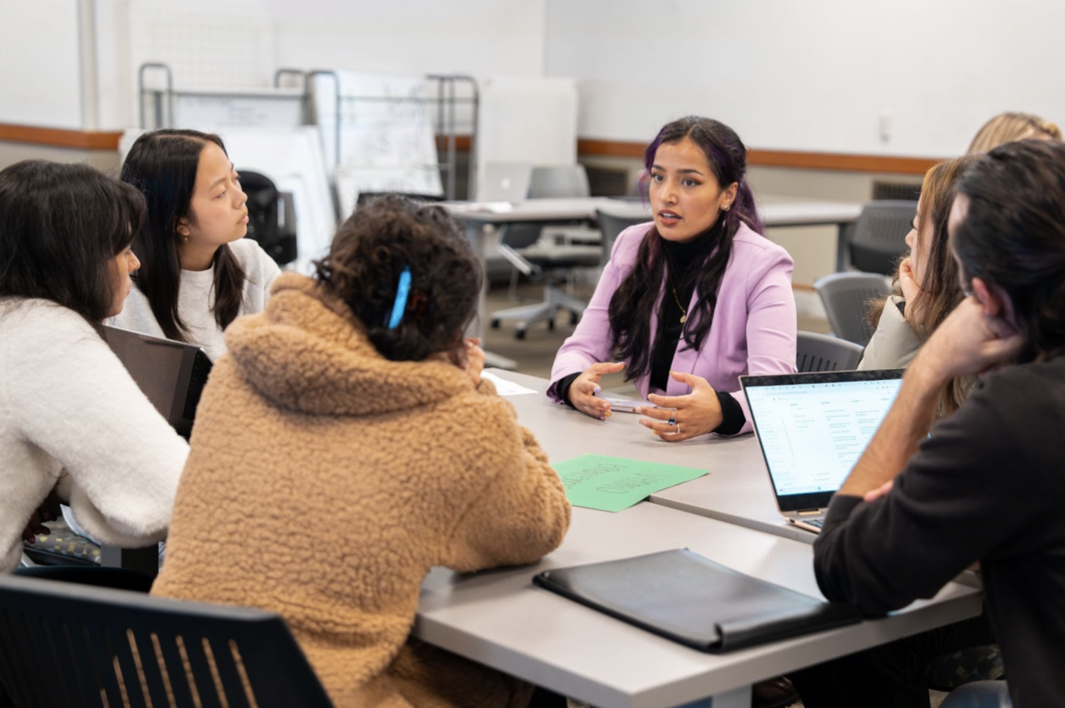 Kashish Juneja sits at a table, speaking to a group of six students who are taking notes on laptops and paper.